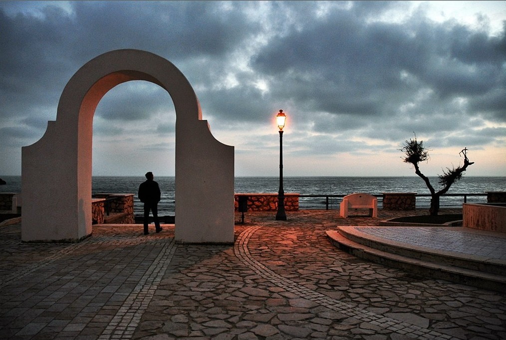 un arco candido incornicia i pensieri mentre il giorno sfuma nel blu profondo del Tirreno. Tra la pietra antica di Sperlonga e un cielo denso di nuvole, la luce solitaria di un lampione accende la sera, sospesa tra il silenzio del borgo e la voce eterna del mare. Questo tramonto è un invito a tornare al momento presente: respirare profondamente, ritrovare il proprio centro e riscoprire la pace che risiede da sempre dentro di noi.