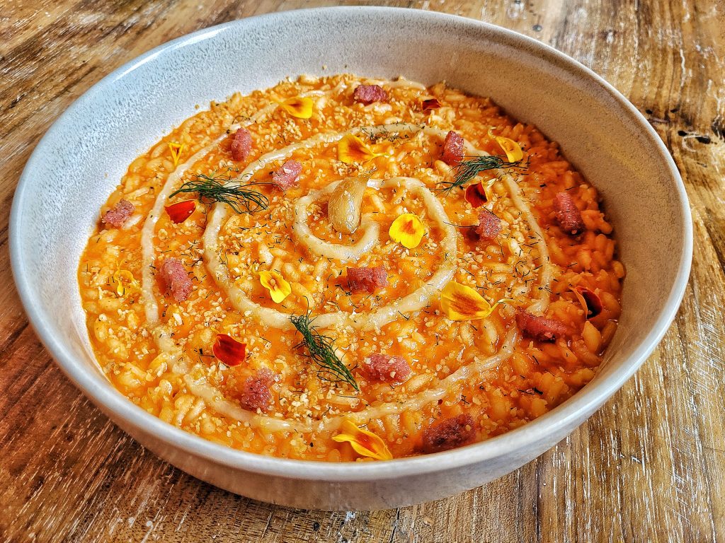 ...l’aglio confit che si fa crema, dolce e profonda, il peperone arrostito accende il piatto di vita. Nulla si spreca: gli scarti dei protagonisti diventano brodo, il pane raffermo si veste di spezie. Un risotto che nutre corpo e coscienza, dove ogni ingrediente ha una seconda possibilità. Il rosso qui non è solo colore: è calore, cura, rispetto. Essenza